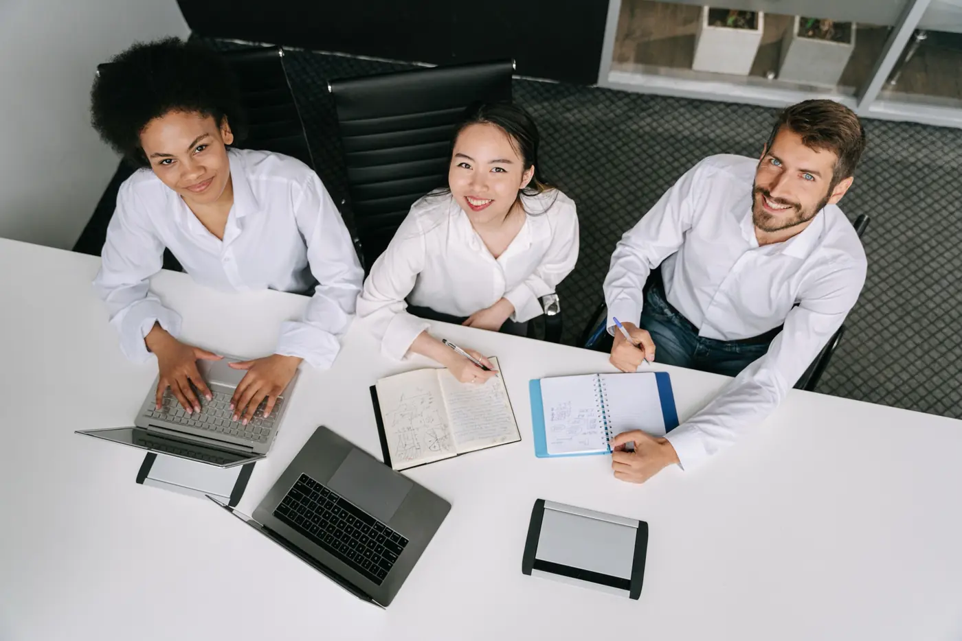 Trois collègues souriants autour d’une table de réunion, avec ordinateurs portables et carnets de notes